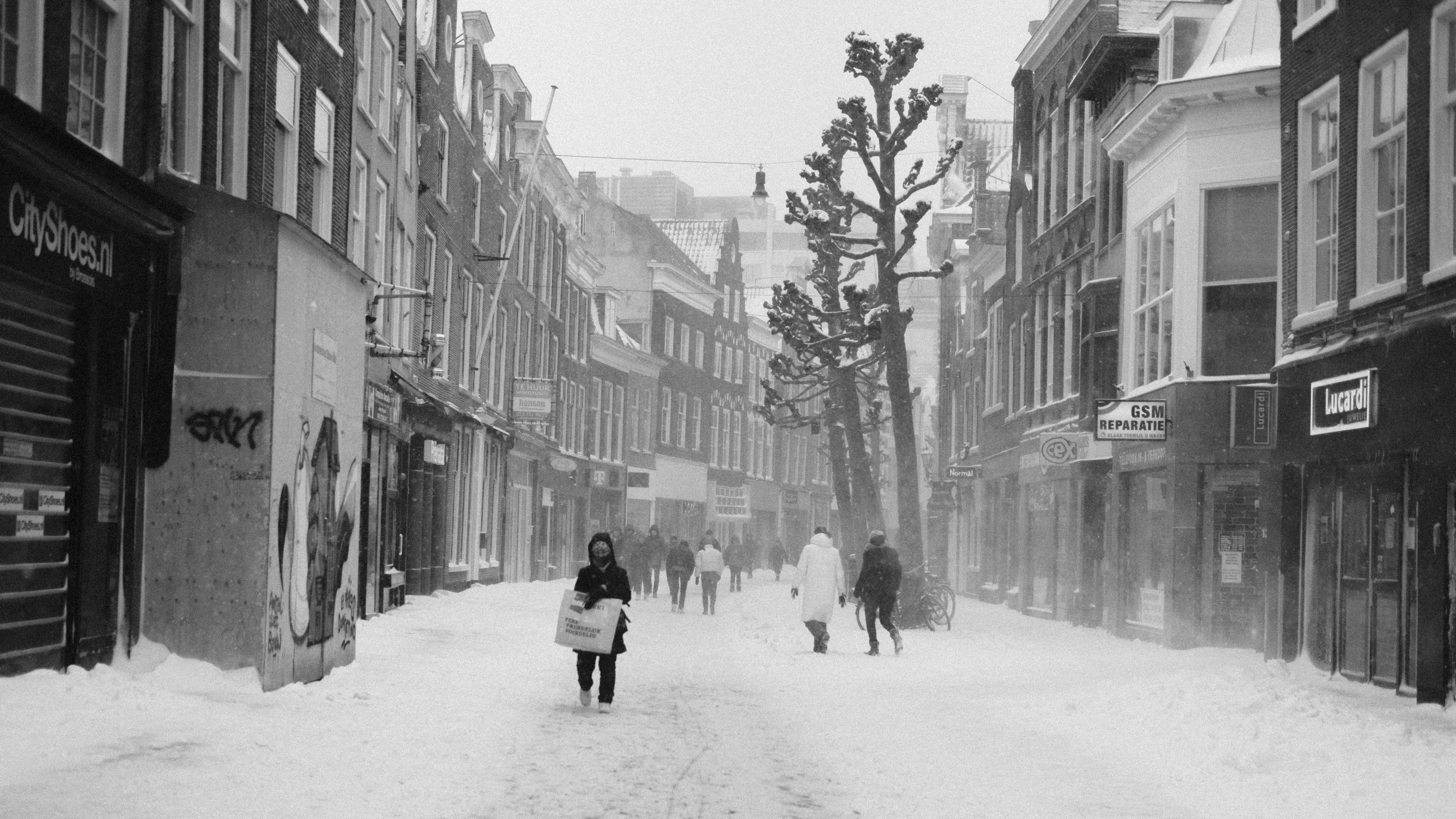 grayscale photo of people walking on snow covered road between buildings